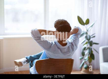 Middle Eastern man looking out of window with hands behind head sitting on chair in living room at home Stock Photo