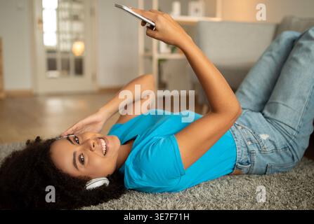 Black woman with curly afro listening to music on headphones and holding smartphone while lying on floor carpet at home Stock Photo