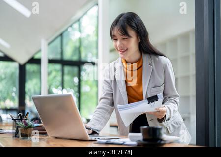 Thai woman working on laptop holding documents standing at table in office during daytime Stock Photo