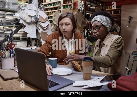 Black and caucasian female tailors holding sandwiches and discussing new design using laptop while sitting at table in workshop Stock Photo