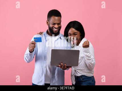 Cheerful Black couple holding laptop and credit card while standing against pink background in studio Stock Photo