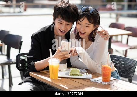 Smiling Chinese couple using smartphone together having lunch while sitting at table in outdoor restaurant on sunny day Stock Photo