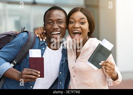 Cheerful Black couple holding passports with plane ticket standing close together outside airport Stock Photo