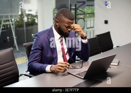 Tired Black businessman in suit massaging his nose bridge holding eyeglasses sitting at table with laptop in office Stock Photo