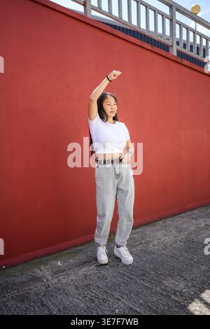 Asian woman in shirt and jeans dancing in front of red wall on city street during daytime Stock Photo