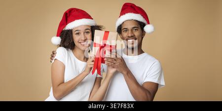 Black couple wearing Santa hats holding gift box with red ribbon while standing together against brown background in studio Stock Photo