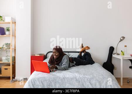 Black Muslim female teenager in brown hijab using laptop lying down on bed at home Stock Photo