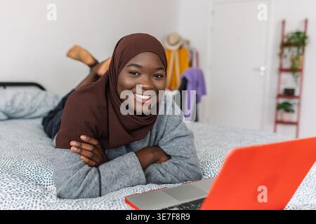 Black Muslim female teenager in brown hijab lying down on bed with laptop at home Stock Photo