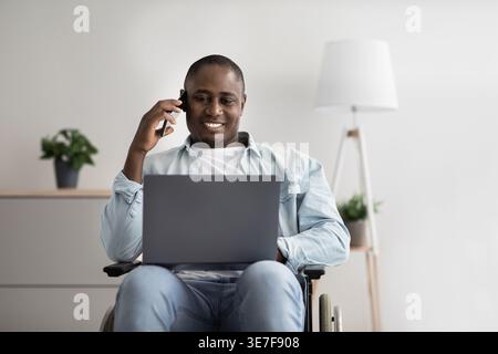 Disabled Black man sitting in wheelchair having phone call and looking at laptop screen working in living room at home Stock Photo