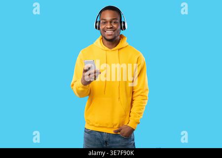 Smiling bearded Black man wearing yellow hoodie listening to music on headphones using smartphone while standing with hand on pocket in front of bl... Stock Photo
