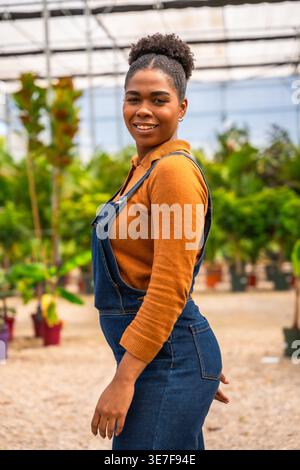 Hispanic female gardener in denim overalls smiling while standing sideways in greenhouse during daytime Stock Photo