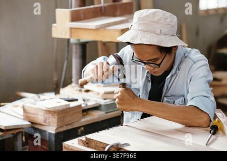 Asian male carpenter wearing bucket hat using chisel and hammer when working with wood board sitting in workshop during daytime Stock Photo
