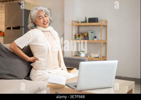 Tired Asian senior woman having back pain sitting on sofa in front of laptop in living room at home Stock Photo