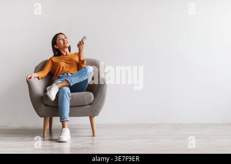 Asian woman using remote adjusting aircon temperature while sitting with crossed legs on chair against white wall in studio Stock Photo