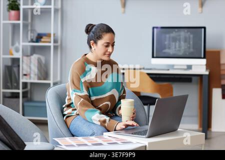 Mixed-race female designer working on laptop while sitting on couch near desk in office Stock Photo