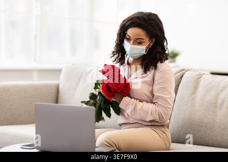 Black woman wearing face mask having video call on laptop holding and showing bouquet of red roses while sitting on sofa in living room at home dur... Stock Photo