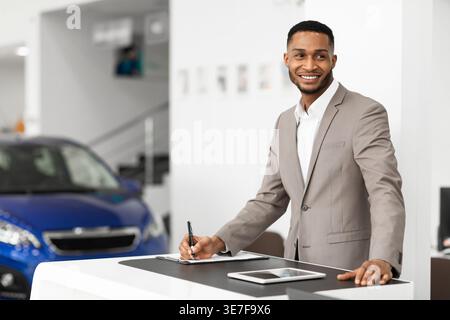 Smiling Black salesman with beard looking away standing at front desk with tablet in car dealership Stock Photo