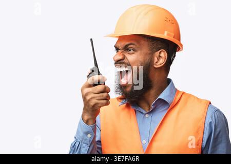 Angry Black male worker wearing safety helmet yelling at walkie-talkie looking away while standing against white background in studio Stock Photo