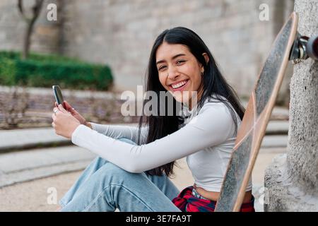 Venezuelan female skater with long black hair holding smartphone sitting on street during daytime Stock Photo