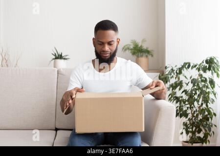 African american millennial man opens cardboard box, received parcel on sofa, free space Stock Photo