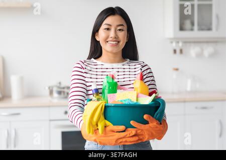 Happy asian woman with cleaning tools posing at kitchen, wearing rubber gloves, young chinese woman holding cleaning tools while house-keeping apartme Stock Photo