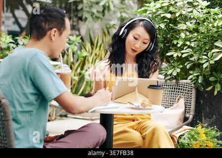 Chinese girlfriend wearing headphones using tablet while sitting at table with her boyfriend drinking iced coffee taking notes in outdoor cafe duri... Stock Photo
