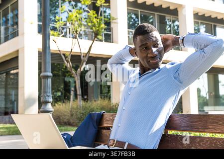 Black businessman stretching arms looking away sitting on wooden bench with laptop in city on sunny day Stock Photo
