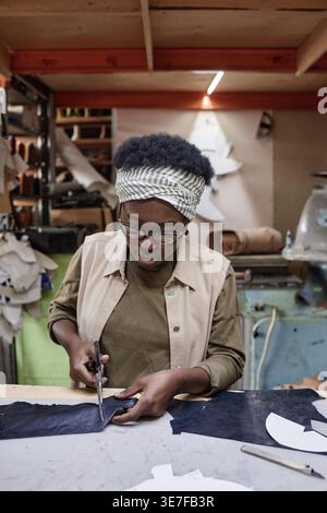 Black female designer wearing eyeglasses cutting leather while sitting at table in workshop Stock Photo