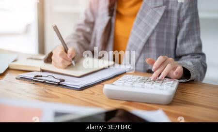 Cropped images of Thai woman taking notes in her notebook while using calculator sitting at table in office Stock Photo