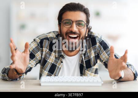 Cheerful Arab man wearing eyeglasses sitting with open hands at table near keyboard in living room at home Stock Photo