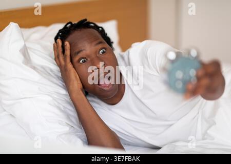 Shocked Black man with dreads looking at alarm clock while lying on bed at home Stock Photo