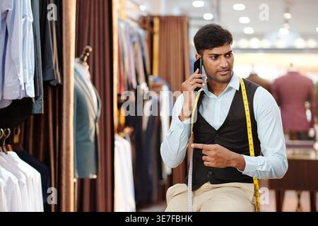 Indian male tailor having phone call looking away while standing and leaning on sofa in atelier Stock Photo