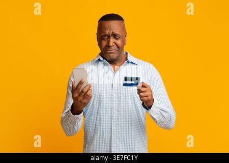 Upset middle-aged Black man holding smartphone and credit card checking his balance while standing against yellow background in studio Stock Photo