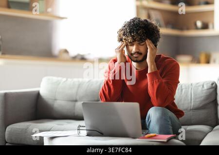 Freelance Stress. Tired Indian Man Suffering Headache While Working On Laptop At Home, Sick Eastern Male Rubbing Temples And Frowning, Having Acute Mi Stock Photo