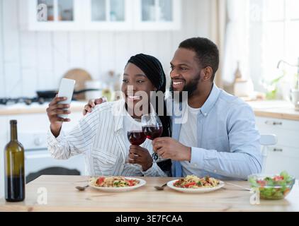 Black couple toasting glasses of wine taking selfie using smartphone sitting at table in kitchen at home Stock Photo