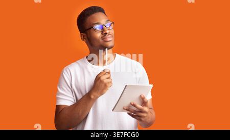 Black man wearing eyeglasses holding notebook and pen on his chin looking away thinking against orange background Stock Photo