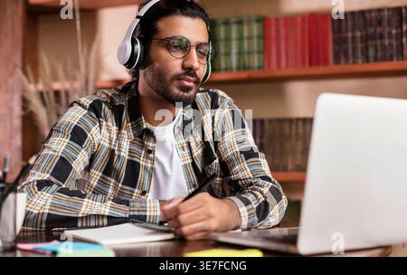 Indian male student in headphones wearing eyeglasses learning online using laptop sitting at table in library Stock Photo
