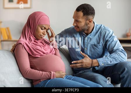 Black Muslim pregnant woman in hijab feeling unwell sitting on sofa and arguing with husband at home Stock Photo