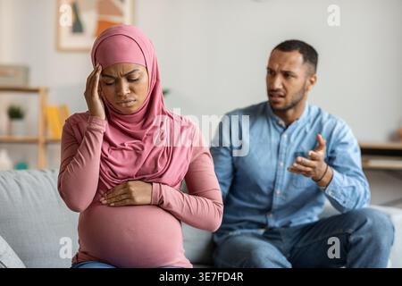 Black Muslim pregnant woman in hijab feeling unwell sitting on sofa and arguing with husband at home Stock Photo