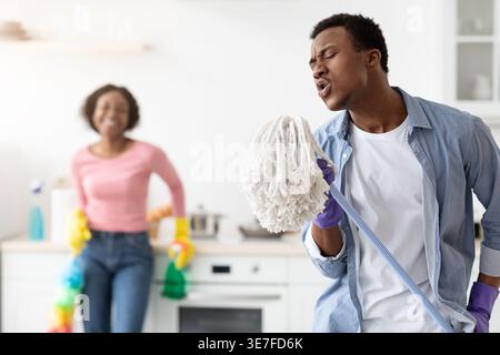 Black man wearing gloves having fun singing using mop handle while cleaning with husband in kitchen Stock Photo