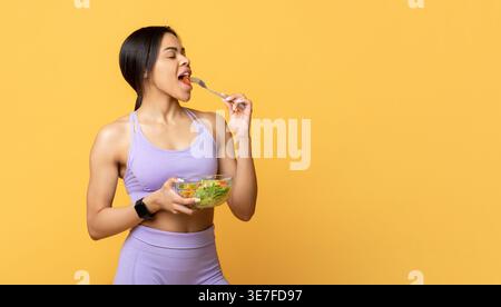 Black woman eating salad while standing with closed eyes against yellow background in studio Stock Photo