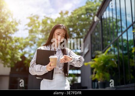 Thai business woman holding clipboard and cup of coffee looking at wristwatch late for work walking near office building in city during daytime Stock Photo