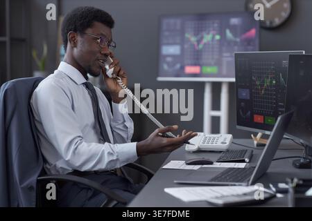 Black male broker talking on the phone with customer while sitting at desk with computers in office Stock Photo