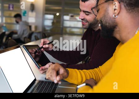 Black man with beard wearing yellow sweater and eyeglasses working with caucasian male colleague discussing project plan on tablet while sitting to... Stock Photo