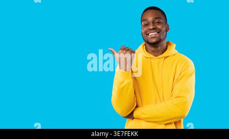 Smiling bearded Black male freelancer wearing hoodie pointing finger sideways while standing in front of blue background in studio Stock Photo