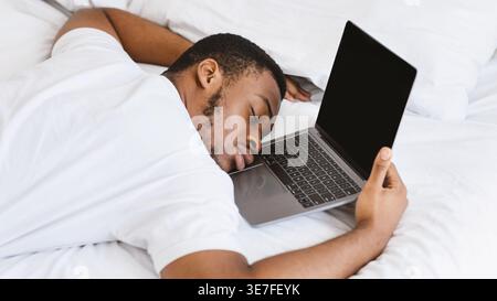 Black man in white shirt fell asleep while using laptop on bed at home Stock Photo