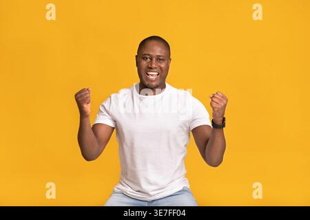 Cheerful  Black man in white standing with clenched fist against orange background in studio Stock Photo