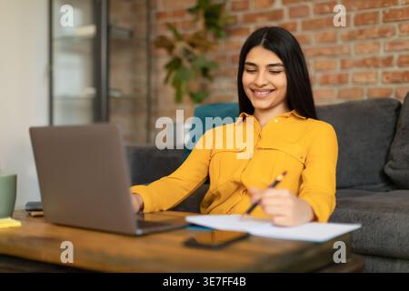 Middle Eastern female student using laptop while writing down notes sitting at desk in living room at home Stock Photo
