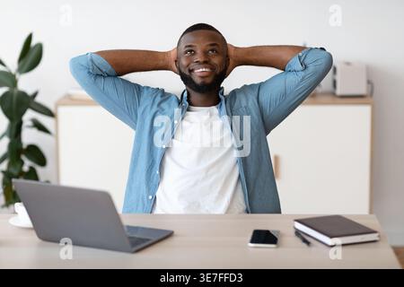 Bearded Black male freelancer wearing blue long-sleeved shirt looking up leaning head head on both hands while sitting at table with laptop and not... Stock Photo