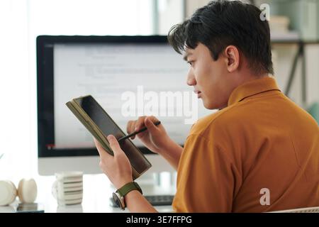 Asian male office worker using tablet sitting at desk with computer in office Stock Photo